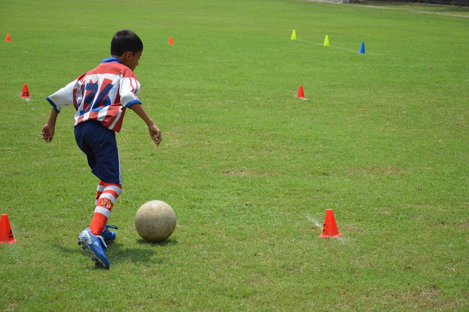 Under-14 boy in a red-and-white striped kit dribbles a football through cones during grassroots training in Kolkata
