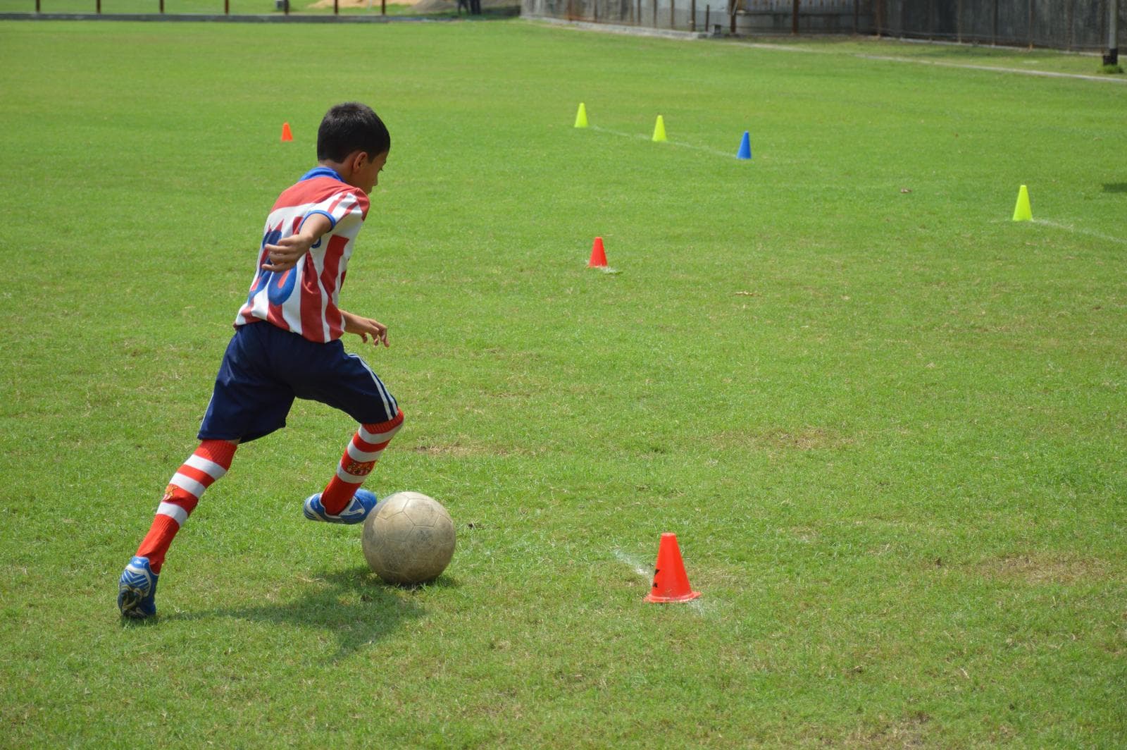 A boy in a red-and-white kit dribbles a football through cones on a grass pitch in Kolkata